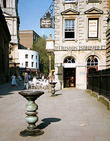 Two ornate metal pillars with large dishes on top in a paved street, with an eighteenth-century stone building behind, upon which can be seen the words